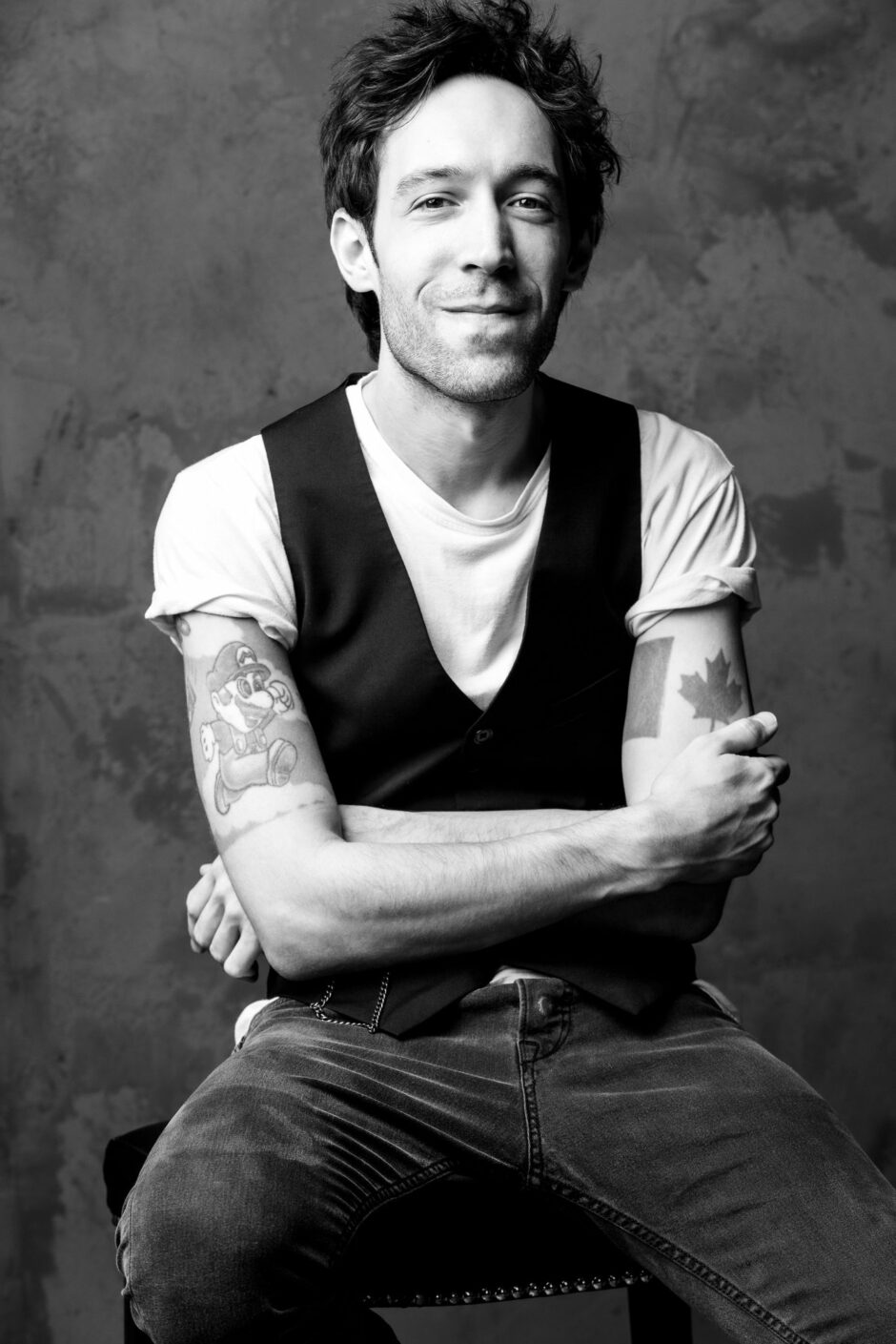A black and white portrait of Creagen Dow sitting with a relaxed, confident expression. He is wearing a sleeveless vest over a white t-shirt, revealing tattoos on both his arms—a cartoon character on his left and a maple leaf on his right. The background is a plain, textured wall.