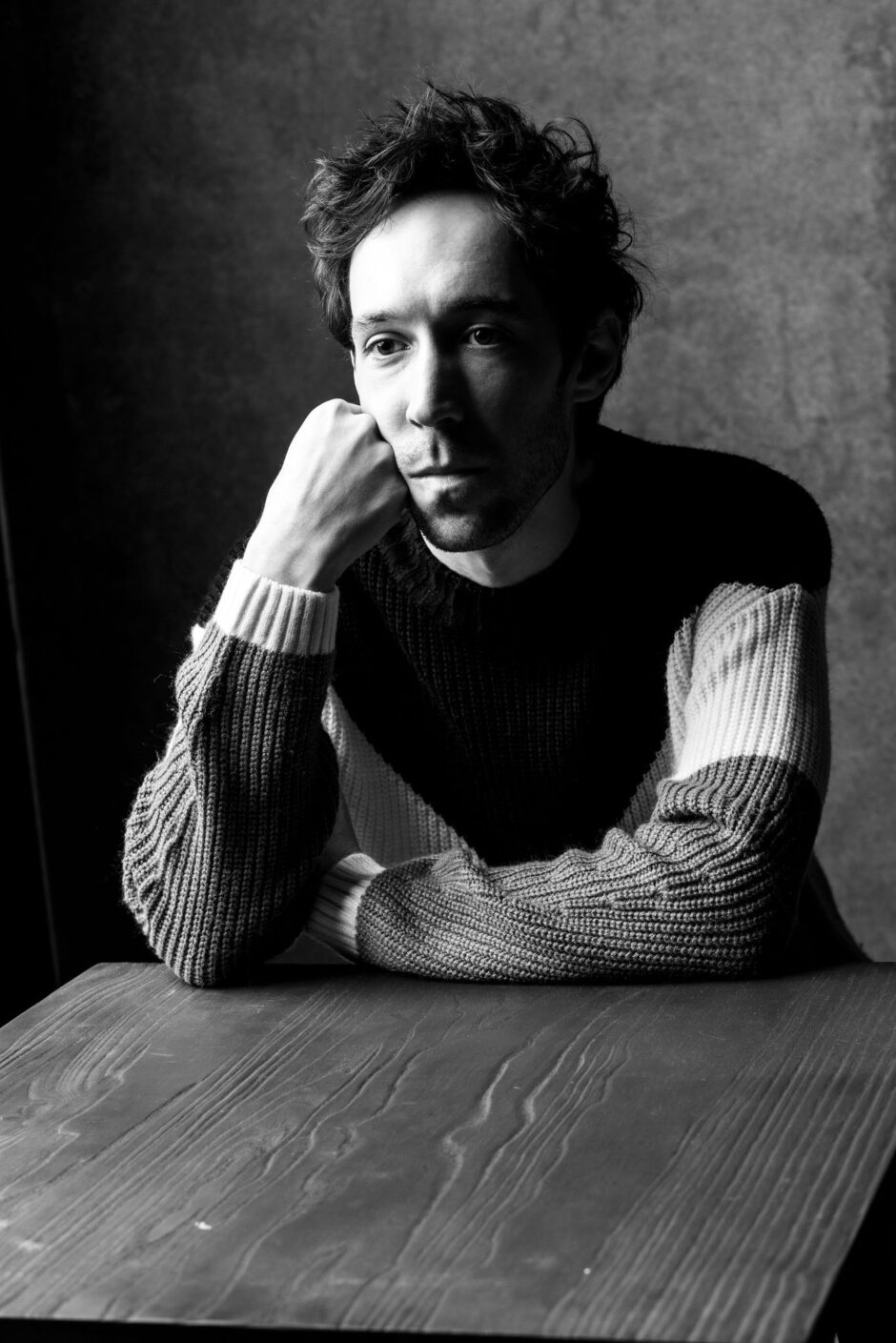 A black and white portrait of Creagen Dow sitting at a wooden table, resting his chin on his fist. He has tousled hair and is wearing a knitted sweater with contrasting colors. His expression is thoughtful and introspective, and the lighting highlights the textures of his sweater and the table's grain. The background is a plain, dark wall.