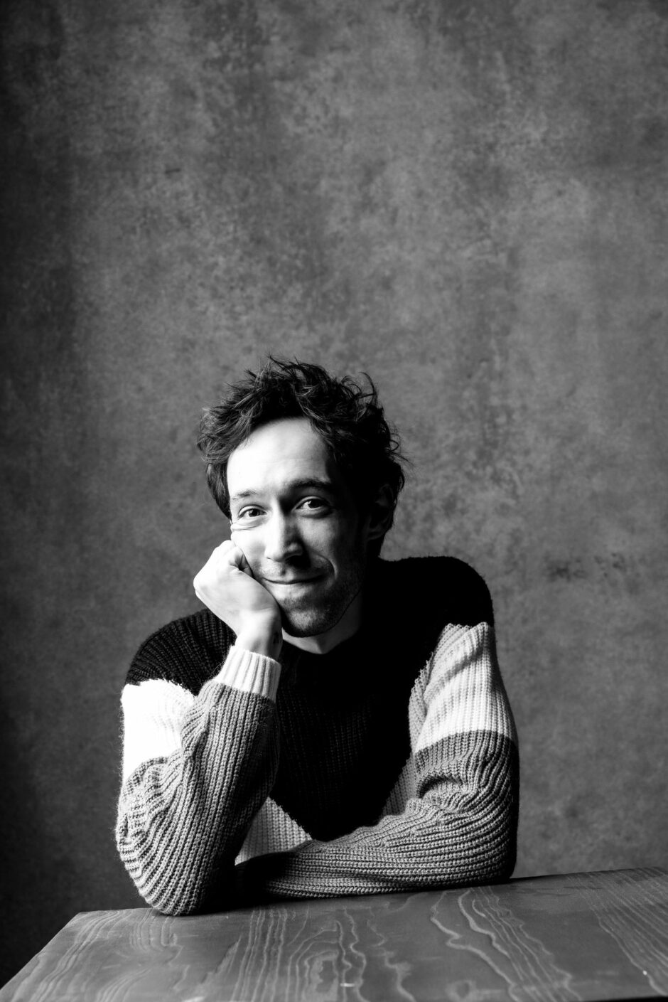 A black and white portrait of Creagen Dow sitting at a wooden table, resting his head on his hand with a slight smile. He is wearing a striped sweater and has tousled hair, creating a casual and relaxed atmosphere against a textured backdrop.